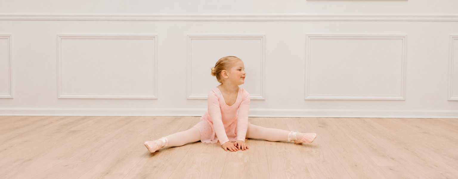 Toddler ballerina in a pink long-sleeve leotard, tights, and ballet shoes sitting in a split stretch on a light wood floor in a bright studio.