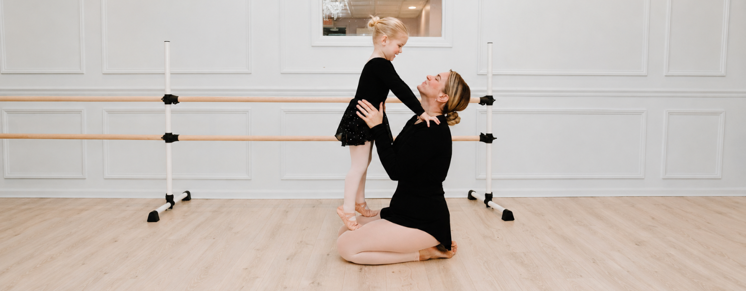 Young ballet student in a black leotard smiling with her teacher during a gentle class moment in a bright dance studio.