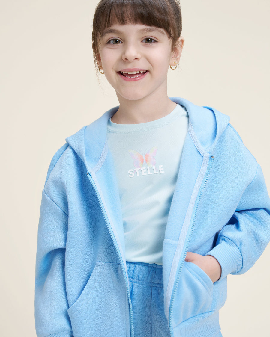 Young girl wearing a light blue hoodie and matching pants with a white shirt underneath on a beige background.