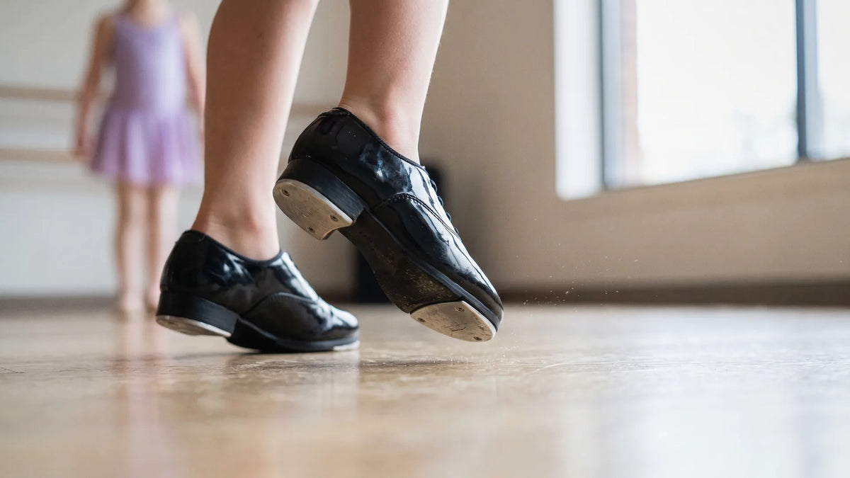 What is tap dance: a young dancer's black tap shoes tapping out a rhythm on a light wood studio floor